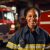 Portrait of a confident African American female firefighter standing in front of the fire truck in her uniform ready to take action